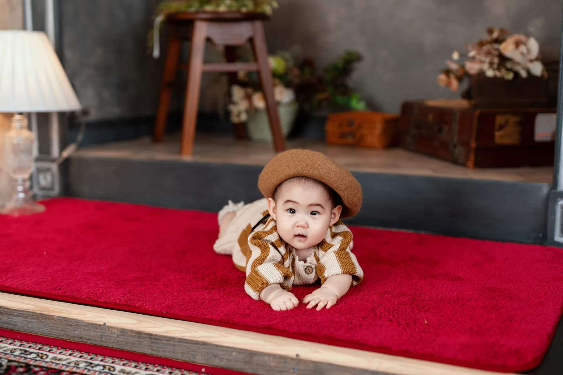 赤いじゅうたんの上でうつぶせになる、ベレー帽の赤ちゃん